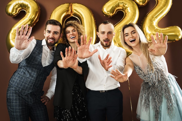 Grupo de gente hermosa fiesta bien vestida que celebra la llegada del año nuevo 2022 con globos dorados sobre fondo marrón