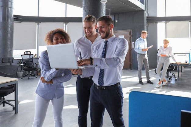 Grupo de gente de negocios moderna está trabajando con ordenador portátil y sonriendo mientras está de pie en el pasillo de la oficina.