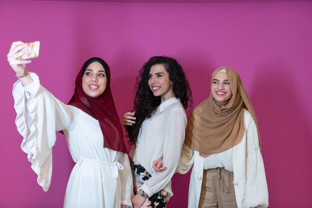 Grupo de hermosas mujeres musulmanas, dos de ellas vestidas a la moda con hiyab usando un teléfono móvil mientras toman una foto selfie aislada en un fondo rosado que representa la tecnología moderna de la moda islámica.