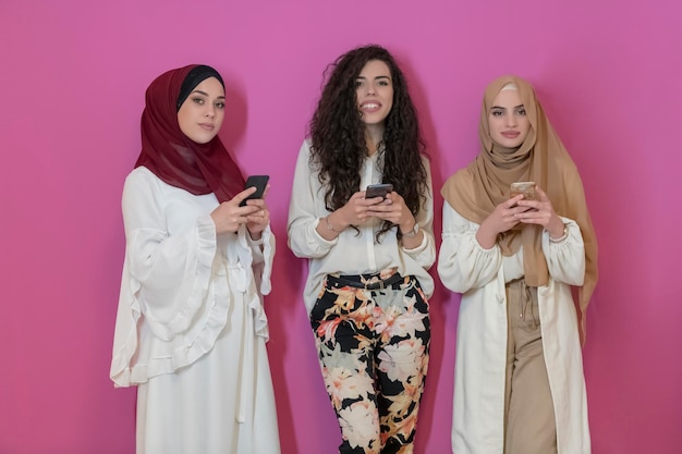 Un grupo de hermosas mujeres musulmanas, dos de ellas con un vestido de moda con hijabs usando teléfonos inteligentes móviles aislados en un fondo rosado, representa la tecnología moderna de la moda islámica y Ramadan Kareem conc.