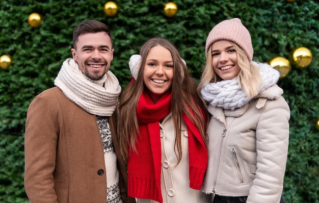 Grupo de hombres y mujeres milenarios positivos en cálidos trajes de invierno sonriendo y mirando mientras están de pie juntos, contra el árbol de Navidad verde decorado con bolas doradas