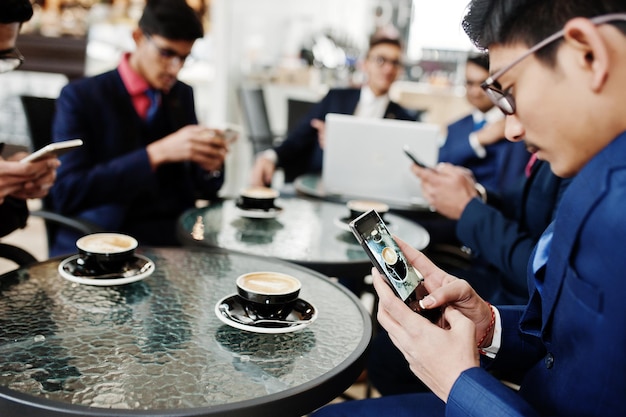 Grupo de hombres de negocios indios en trajes sentados en la oficina en un café con mensajes de texto portátiles en los teléfonos y haciendo fotos de café