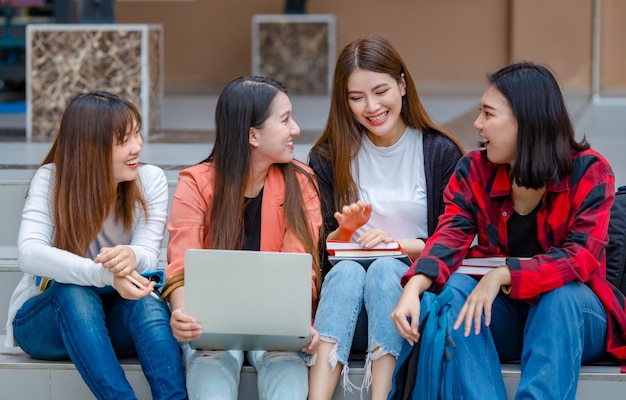 Grupo de inteligentes estudiantes asiáticas con laptop y cuadernos sentados en el campus universitario e investigando juntos mientras miran a la cámara. Compañero íntimo de adolescentes en la universidad.