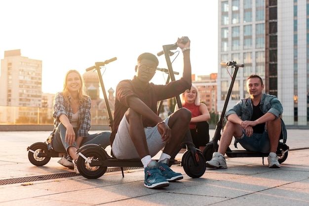 Grupo inter-racial de jovens amigos sentados com patinetes elétricos na cidade e sorrindo