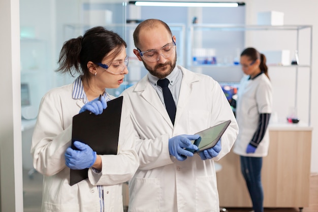Grupo de investigadores médicos que discuten sobre el desarrollo de vacunas, de pie en un laboratorio equipado apuntando a una tableta y tomando notas