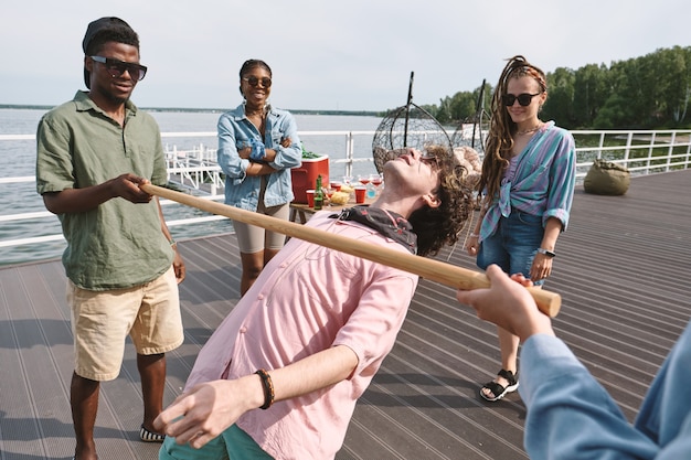 Grupo de jóvenes alegres jugando al juego de limbo en el muelle junto al lago mientras disfrutan de una pequeña fiesta