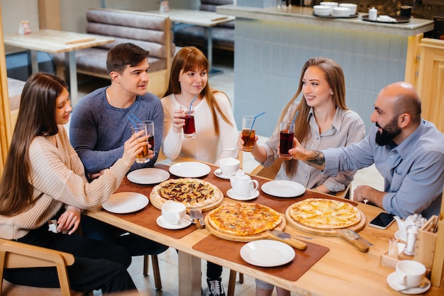 Un grupo de jóvenes amigos alegres está sentado en un café hablando y comiendo pizza. Almuerzo en la pizzería.