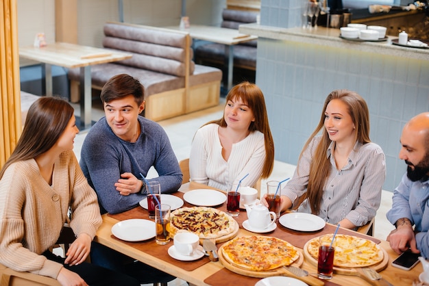 Un grupo de jóvenes amigos alegres está sentado en un café hablando y comiendo pizza. Almuerzo en la pizzería.