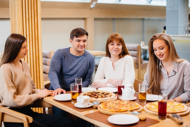 Un grupo de jóvenes amigos alegres está sentado en un café hablando y comiendo pizza