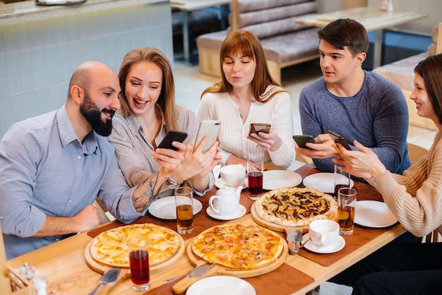 Un grupo de jóvenes amigos alegres está sentado en un café hablando y tomando selfies por teléfono. Almuerzo en la pizzería.