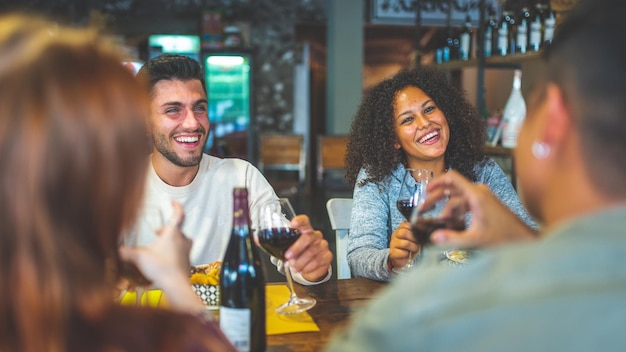 Grupo de jóvenes amigos felices almorzando en el restaurante, bebiendo vino tinto y brindando, concéntrese en la morena colombiana, filtro cálido