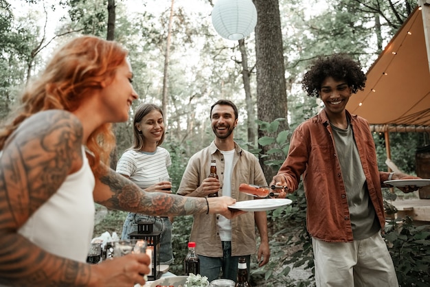 Grupo de jóvenes amigos felices haciendo barbacoa en el bosque en glamping. Hombre africano asar salchichas pasando comida del campamento a su amigo, riendo, divirtiéndose en un picnic en el campo, asando a la parrilla