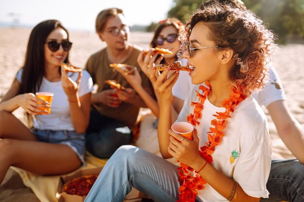 Grupo de jóvenes amigos haciendo un picnic comiendo pizza brindando con beerus en la playa