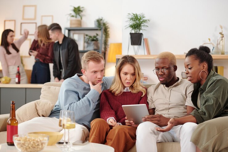 Foto grupo de jóvenes amigos interculturales alegres sentados en fila en el sofá y discutiendo fotos en las redes sociales en la pantalla del dispositivo móvil