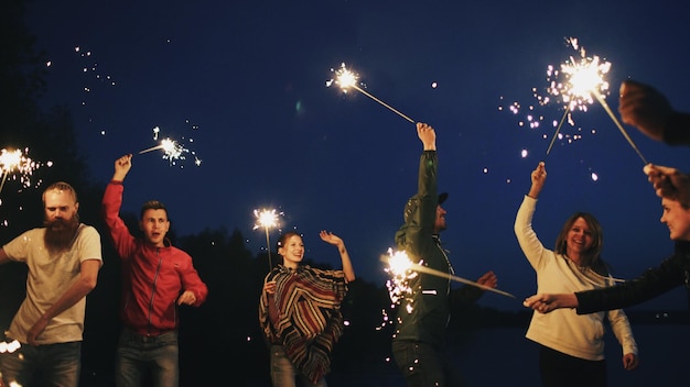 Grupo de jóvenes amigos que tienen una fiesta en la playa bailando y celebrando con bengalas al atardecer