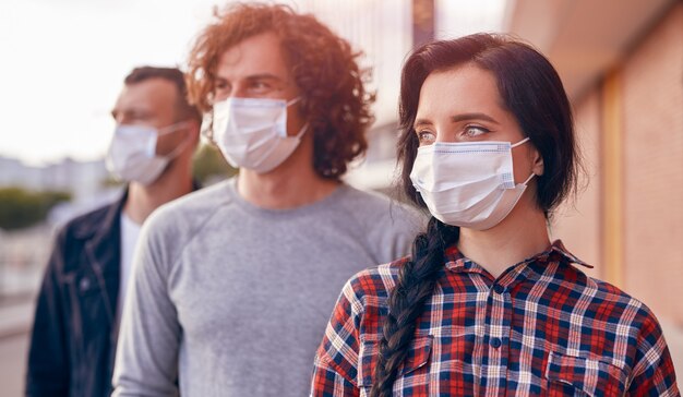 Grupo de jóvenes amigos con ropa casual y máscaras médicas de pie en las calles de la ciudad y mirando hacia el futuro durante la pandemia de coronavirus