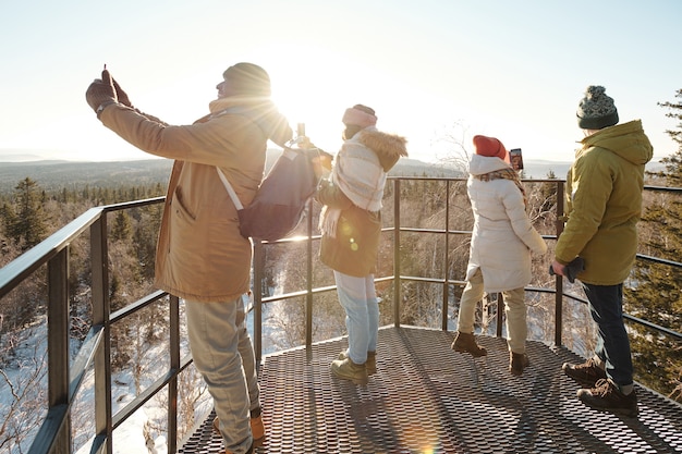 Grupo de jóvenes amigos en ropa de invierno tomando fotos del majestuoso paisaje de montañas y bosques cubiertos de nieve contra el sol que brilla en el cielo azul