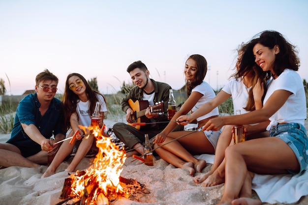 Grupo de jóvenes amigos sentados en la playa y freír salchichas Un hombre toca la guitarra Tiempo de camping