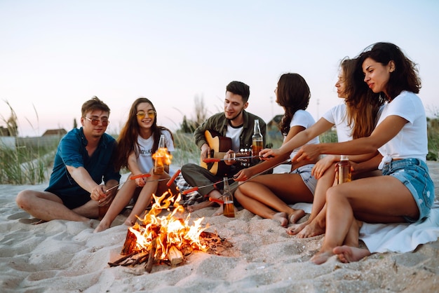 Grupo de jóvenes amigos sentados en la playa y freír salchichas Un hombre toca la guitarra Tiempo de camping