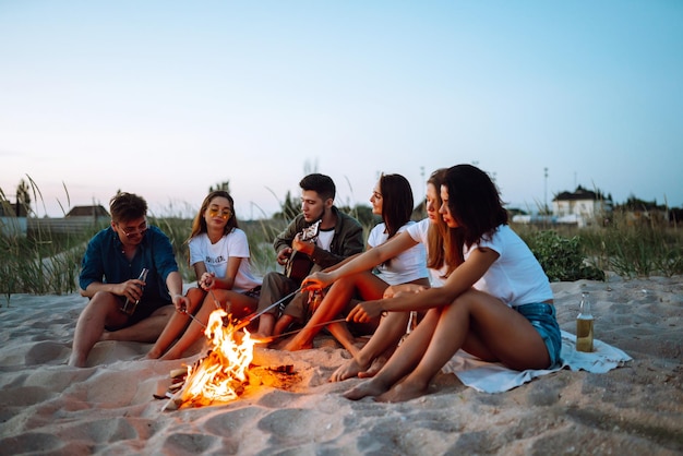 Grupo de jóvenes amigos sentados en la playa y freír salchichas Un hombre toca la guitarra Tiempo de camping