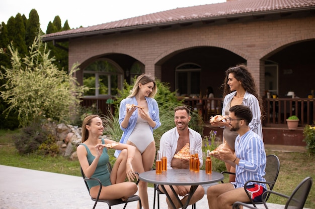 Grupo de jóvenes animando con sidra y comiendo pizza junto a la piscina en el jardín