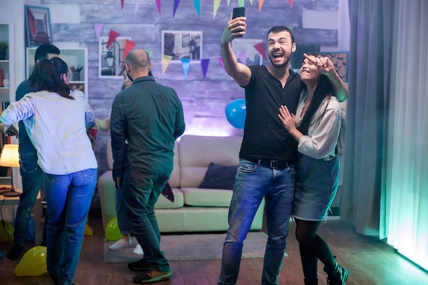 Grupo de jóvenes bailando en una fiesta mientras el hombre y la mujer se toman un selfie.