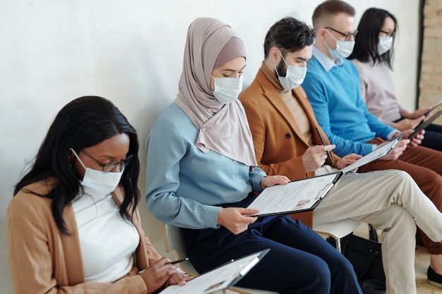Grupo de jóvenes candidatos de trabajo multiétnicos en máscaras sentados en fila y examinando currículum antes de la entrevista