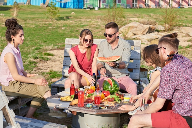 Grupo de jóvenes caucásicos sentados a la mesa al aire libre, comiendo sandía, descansando y charlando en las cálidas noches de verano