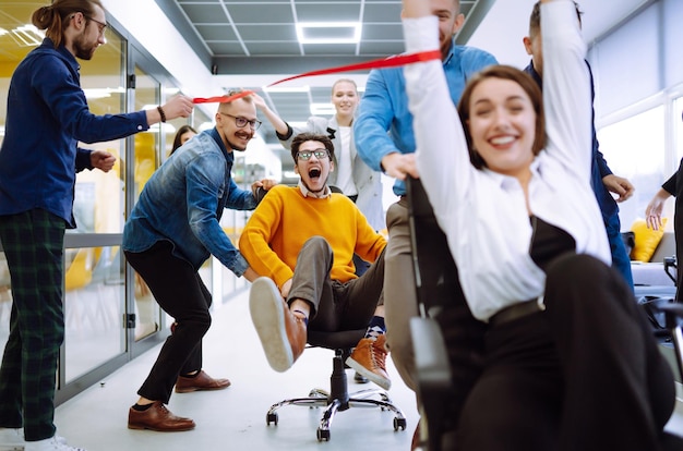 Grupo de jóvenes colegas divirtiéndose juntos montando en sillas en la oficina