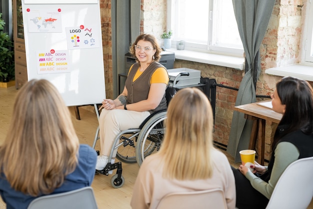 Grupo de jóvenes diseñadoras sentadas en sillas en fila frente a su colega en silla de ruedas haciendo presentación de nuevas ideas en pizarra