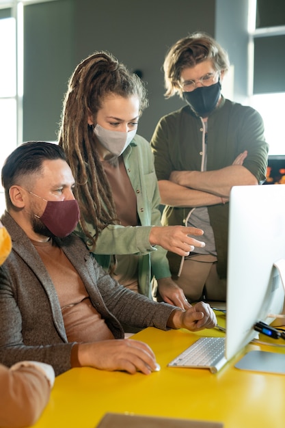 Grupo de jóvenes diseñadores creativos con máscaras protectoras mirando a través de imágenes gráficas en la pantalla del ordenador mientras se discuten nuevas ideas