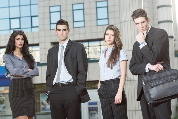 Grupo de jóvenes empresarios exitosos parados frente al edificio de oficinas vestidos con trajes. Con una expresión seria en sus rostros mirando a la cámara.