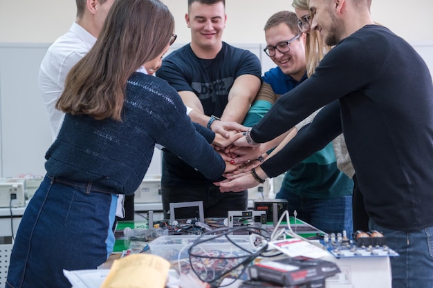 Grupo de jóvenes estudiantes en el aula de electrónica celebrando un proyecto terminado con éxito manteniendo sus manos juntas, concepto de educación y tecnología