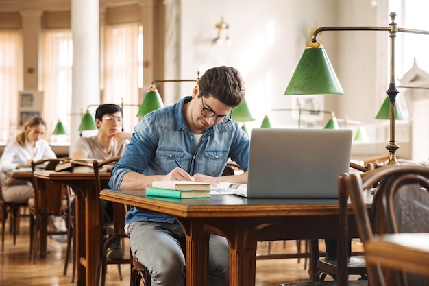 Grupo de jóvenes estudiantes inteligentes que estudian duro en la biblioteca, usando una computadora portátil, leyendo libros