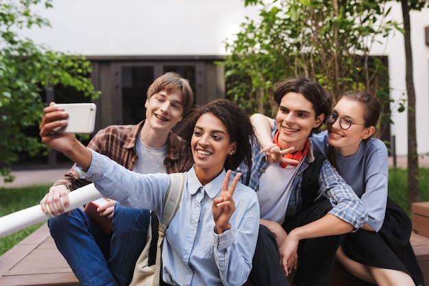 Grupo de jóvenes estudiantes sonrientes sentados y tomando lindas fotos en el teléfono celular mientras pasan tiempo juntos en el patio de la universidad