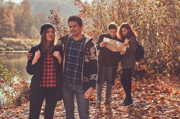 Grupo de jóvenes excursionistas en el bosque de otoño. Adorable pareja abrazándose en primer plano y pareja mirando el mapa y planeando una caminata en el fondo.