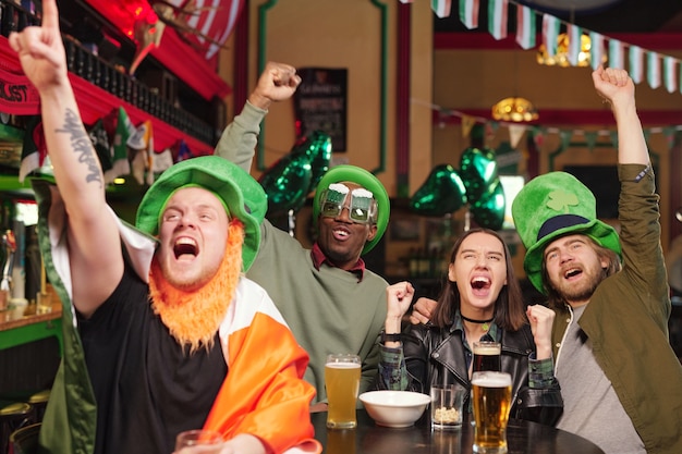 Grupo de jóvenes fanáticos del fútbol intercultural emocionados con sombreros verdes y ropa informal que expresan su triunfo durante la transmisión de fútbol en el pub