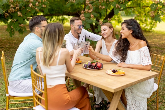 Grupo de jóvenes felices animando con limonada fresca y comiendo frutas en el jardín