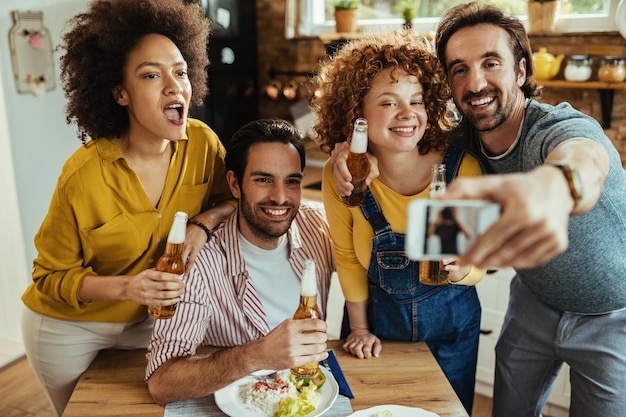 Grupo de jóvenes felices divirtiéndose mientras toman selfie y beben cerveza en el comedor.