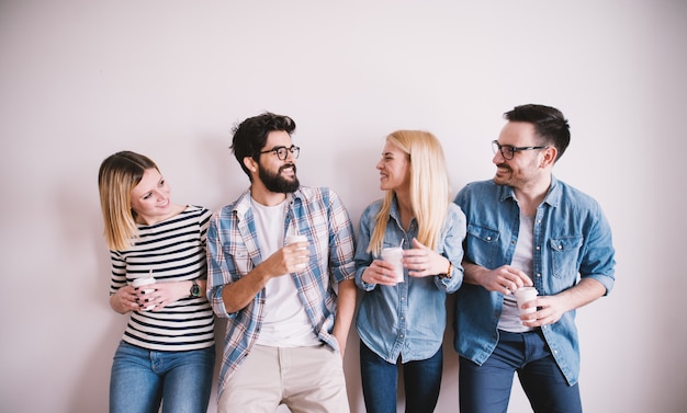 Grupo de jóvenes felices con estilo feliz apoyado contra la pared y hablando mientras bebe café en la taza de papel.