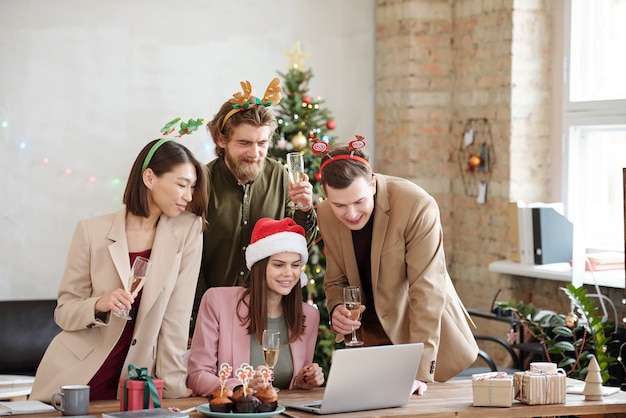 Grupo de jóvenes gerentes alegres con bebidas mirando la pantalla del portátil mientras brindan y felicitan a sus colegas en la fiesta de Navidad en la oficina