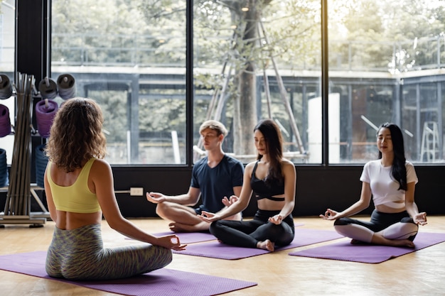 Grupo de jóvenes haciendo yoga sobre una estera de yoga con un entrenador gradualmente enseñando.