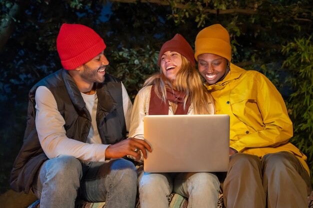 Grupo de jóvenes interraciales que usan una computadora portátil mientras acampan en el campo por la noche hablando por video