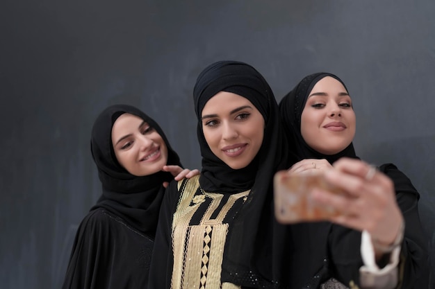 Foto grupo de jóvenes mujeres musulmanas hermosas vestidas de moda con hiyab usando un smartphone mientras toman una foto selfie frente a un fondo negro. foto de alta calidad