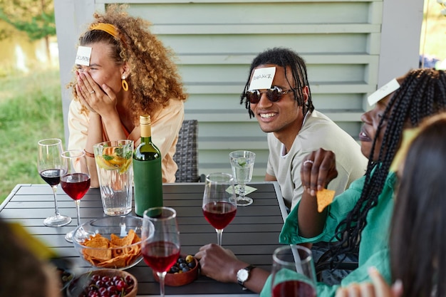 Grupo de jóvenes riendo y jugando a adivinar quién juega mientras se sientan en la mesa al aire libre en verano