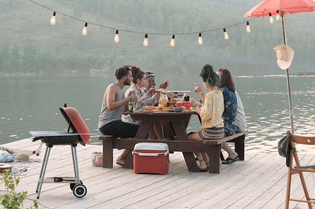 Grupo de jóvenes sentados a la mesa y almorzar durante un picnic en un muelle al aire libre