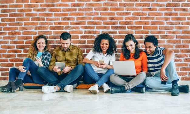 Grupo de jóvenes con teléfonos inteligentes, tabletas digitales y portátiles sentados en fila frente a una pared de ladrillo