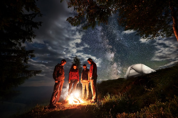 Grupo de jóvenes turistas de pie alrededor del fuego cerca de árboles y carpas