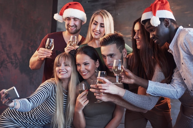 Grupo jovens bonitos fazendo selfie na festa de ano novo, melhores amigas meninas e meninos juntos se divertindo, posando de pessoas em estilo de vida emocional. Chapéus de Papai Noel e taças de champanhe nas mãos