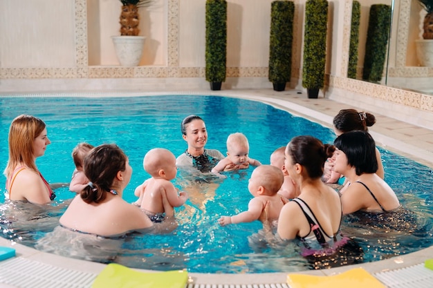 Un grupo de madres con sus hijos pequeños en una clase de natación infantil con un entrenador.